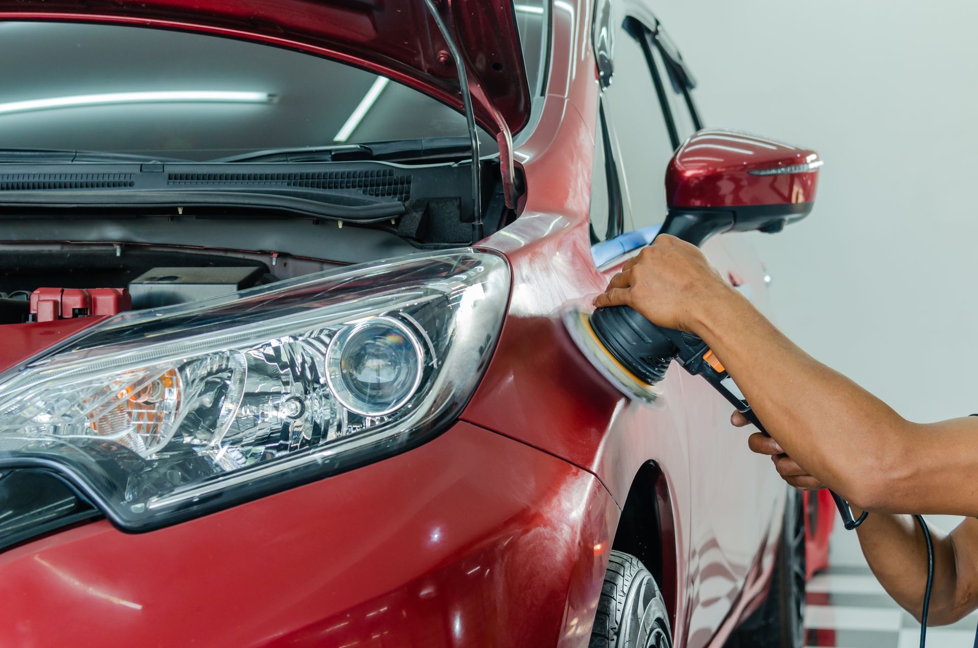 Person using a polisher on a red car in a well-lit shop, near the headlight and side mirror.