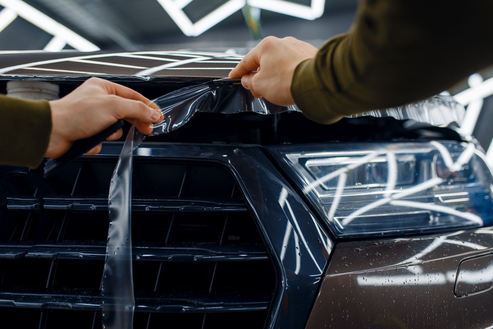 Hands applying clear protective film to a car's front grill.