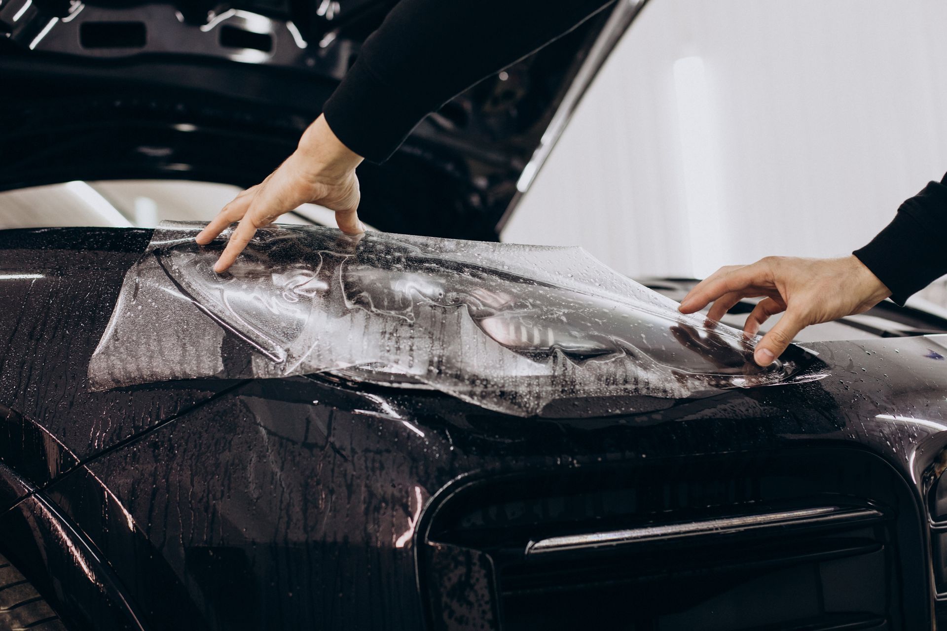 Hands applying protective film to a car headlight. The car is black, the hood is open.