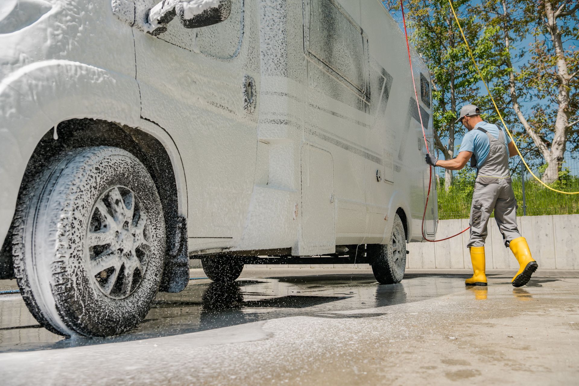 Person washing a white RV with foam and a hose, on a concrete surface.