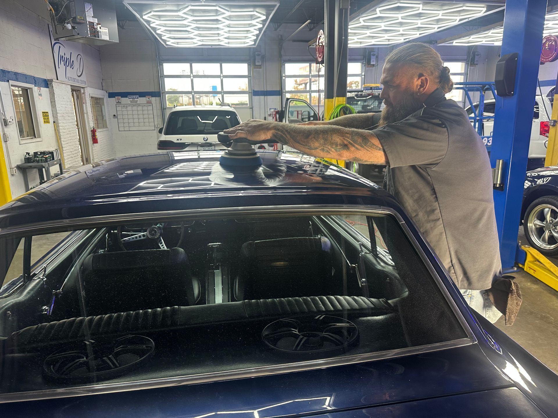 A man polishes the roof of a blue classic car inside a well-lit auto shop.