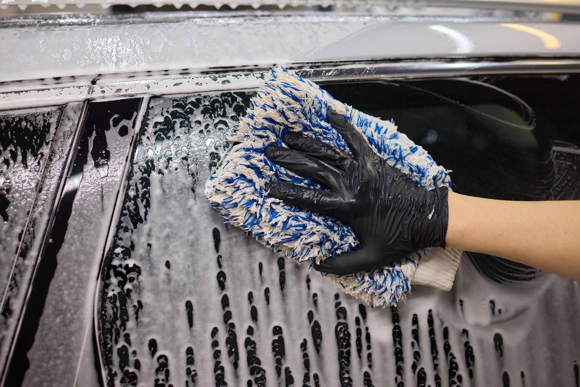 Hand wearing a black glove wiping car window with a soapy microfiber mitt.
