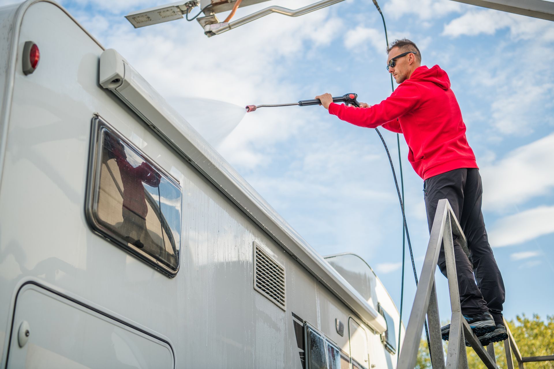 Man in red hoodie power washing the roof of a white RV, standing on a metal platform outdoors.