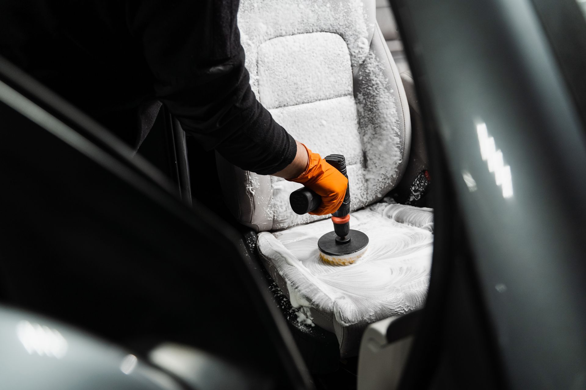Person cleaning a car seat with a power scrubber and orange gloves.