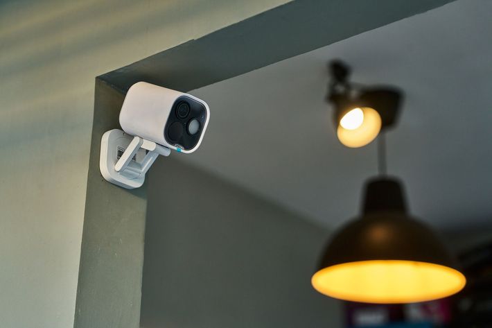 Telecamera di sicurezza bianca montata su una parete grigia, puntata verso una stanza con una lampada sospesa.