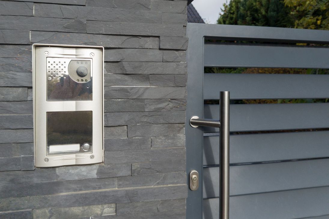 Citofono su un muro di pietra grigio scuro accanto a un moderno cancello grigio con maniglia argentata e serratura a chiave.