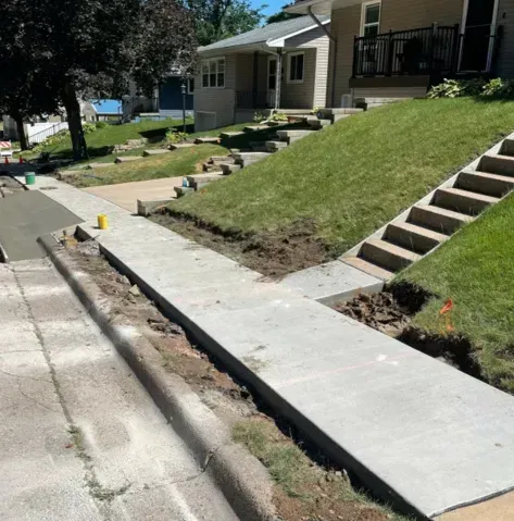 Damaged sidewalk with a curb cut beside a sloped lawn and house steps on a residential street