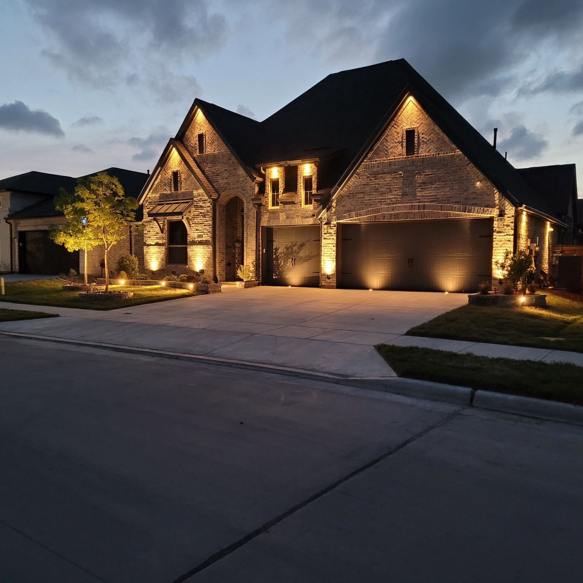 A two-story house with outdoor lighting illuminating the brick facade, driveway, and landscape lighting at dusk.