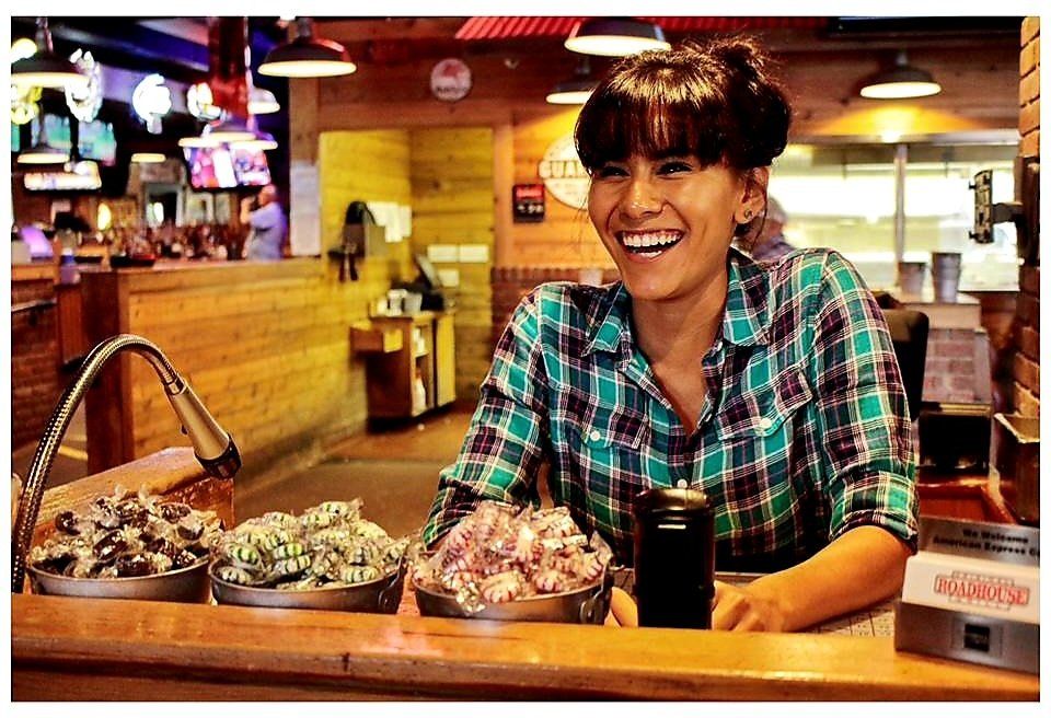 A woman in a plaid shirt is smiling behind a counter in a restaurant.