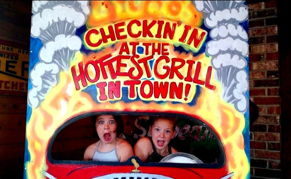 Two girls are looking out of a car window with a sign that says check in at the hottest grill in town