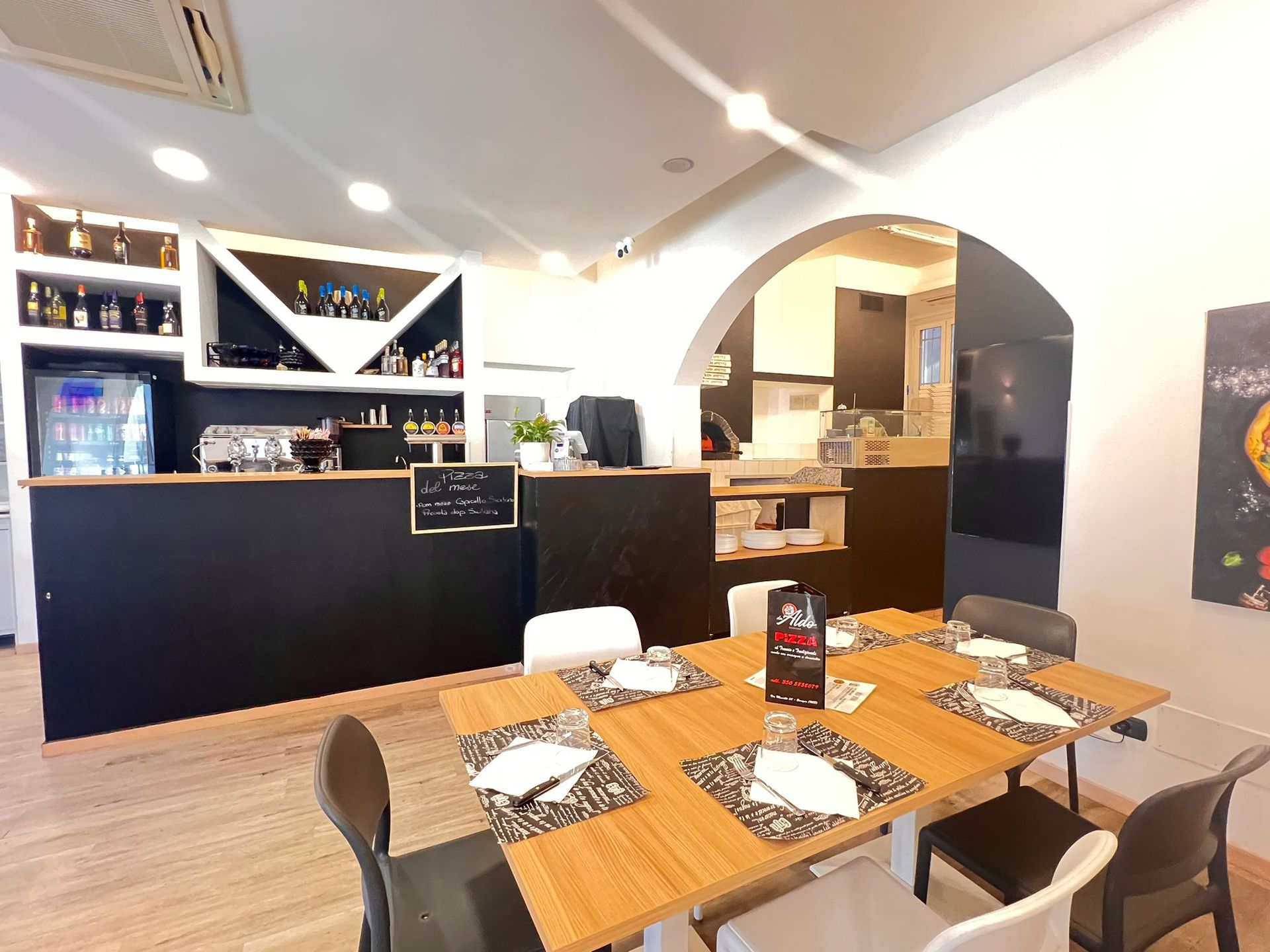 Interno di un ristorante: bar con scaffali pieni di liquori, tavoli apparecchiati per mangiare e un ingresso ad arco che conduce alla cucina.