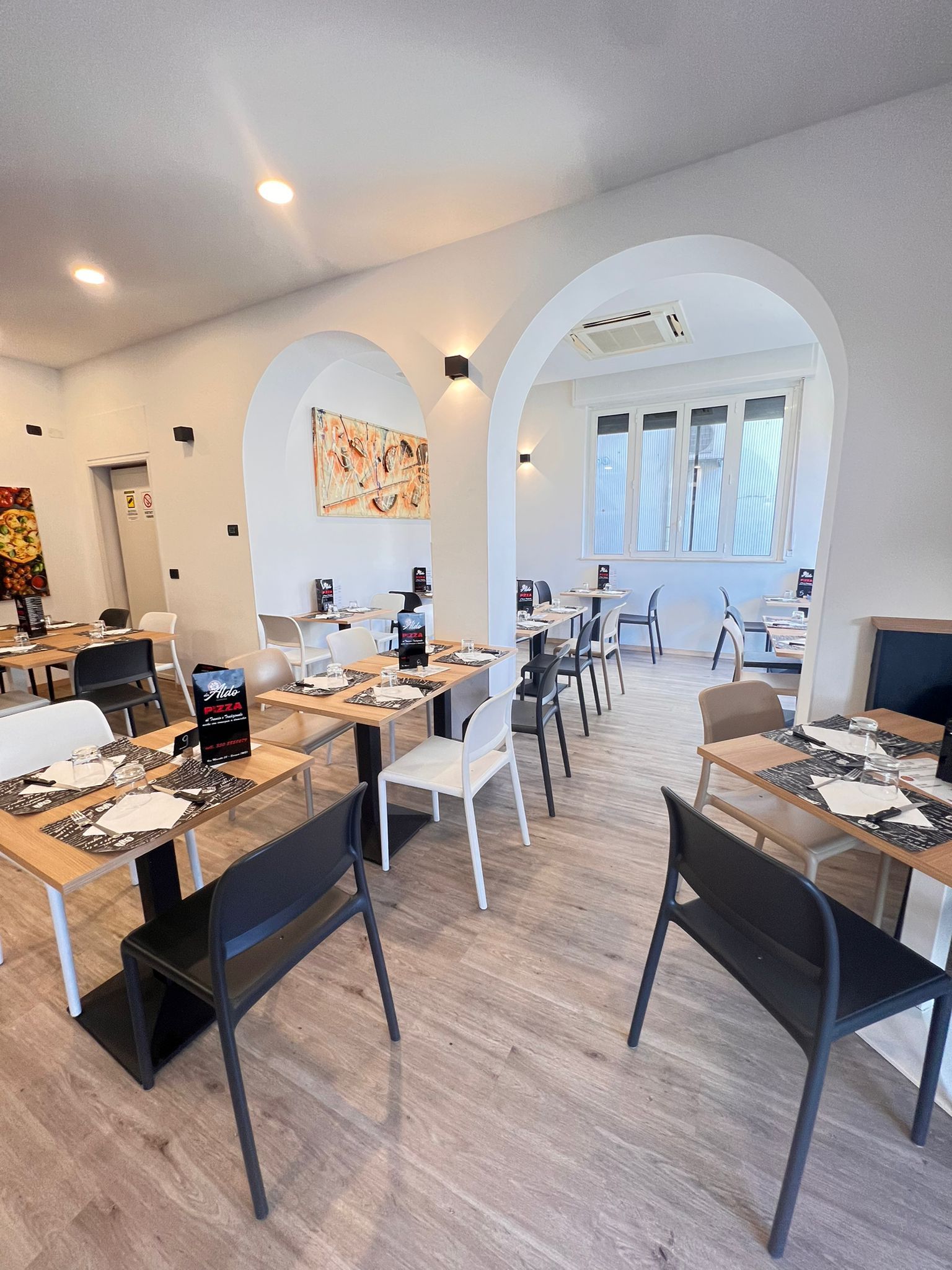 Interno del ristorante con tavoli apparecchiati per la cena, pareti bianche, porte ad arco e pavimenti in legno chiaro.