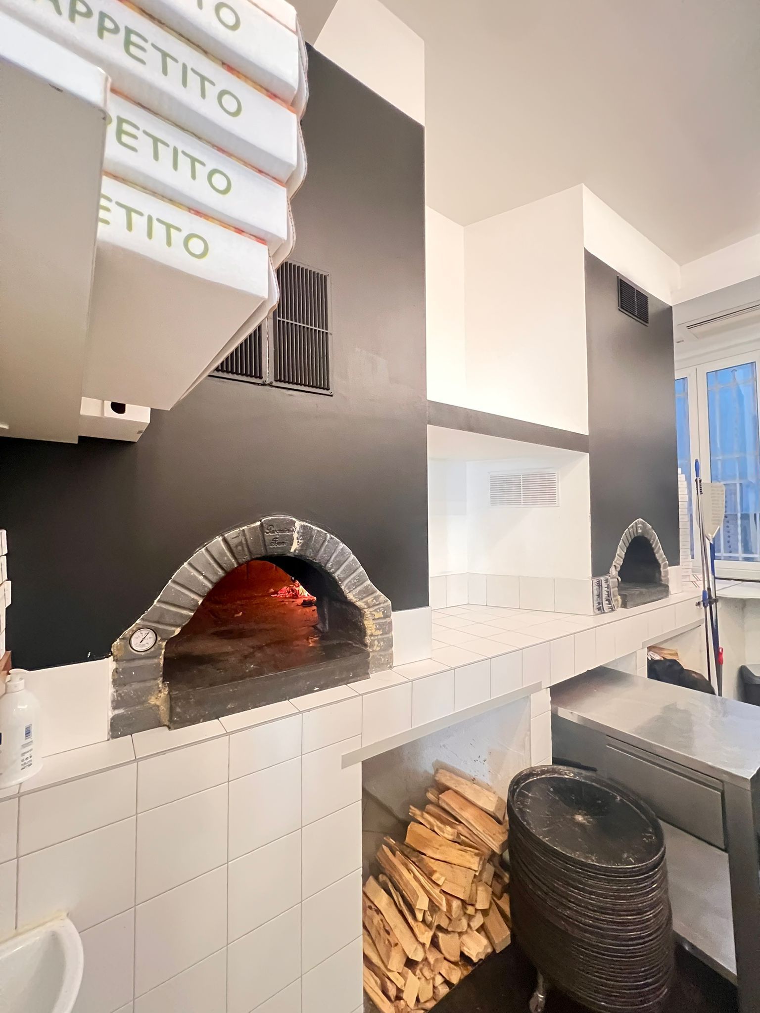Forni per pizza con archi in mattoni, integrati in pareti nere, bancone bianco, legno, scatole per pizza.