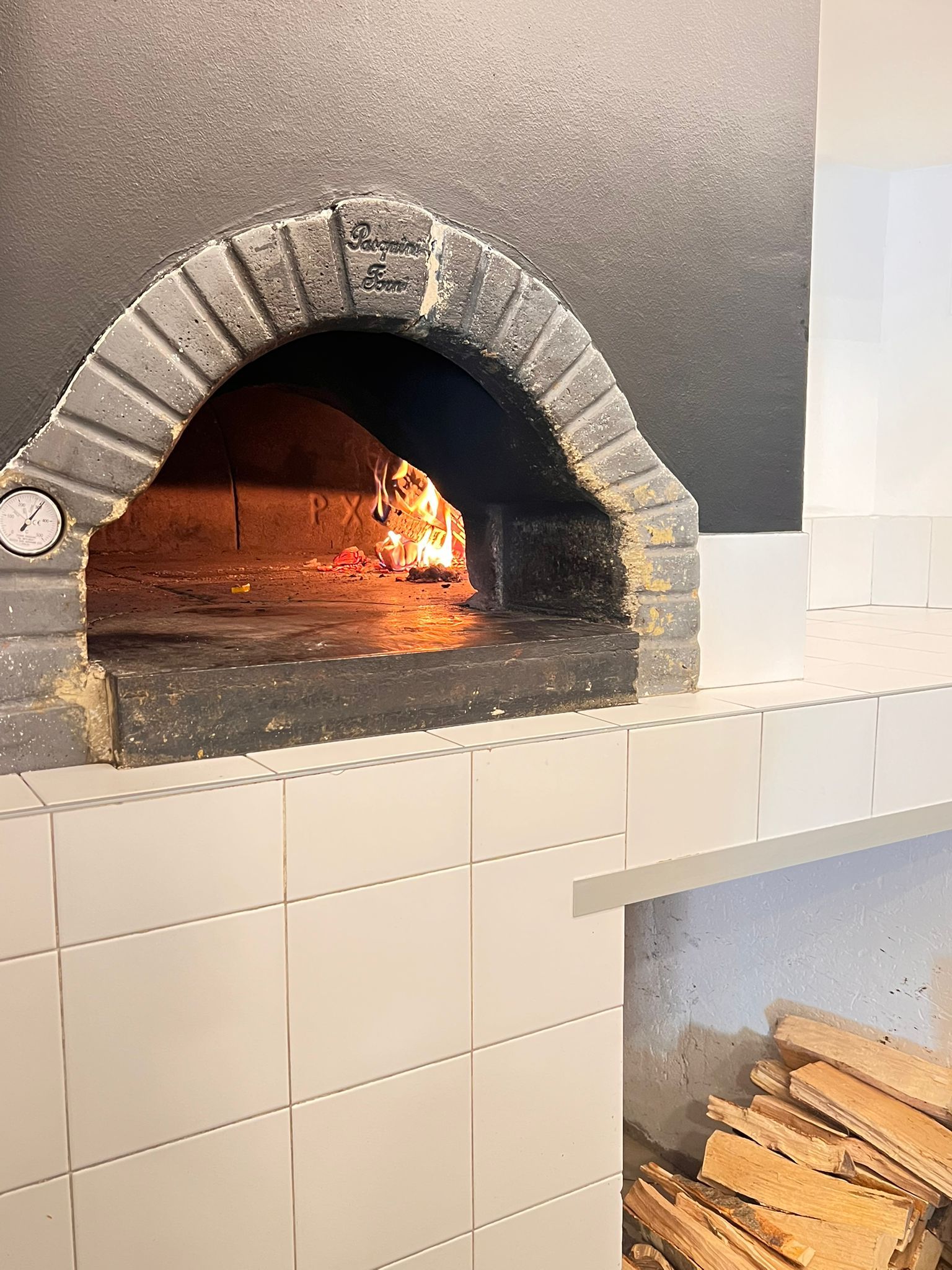 Forno a legna per pizza con fiamme interne. Il forno ha un arco in mattoni ed è circondato da piastrelle bianche.