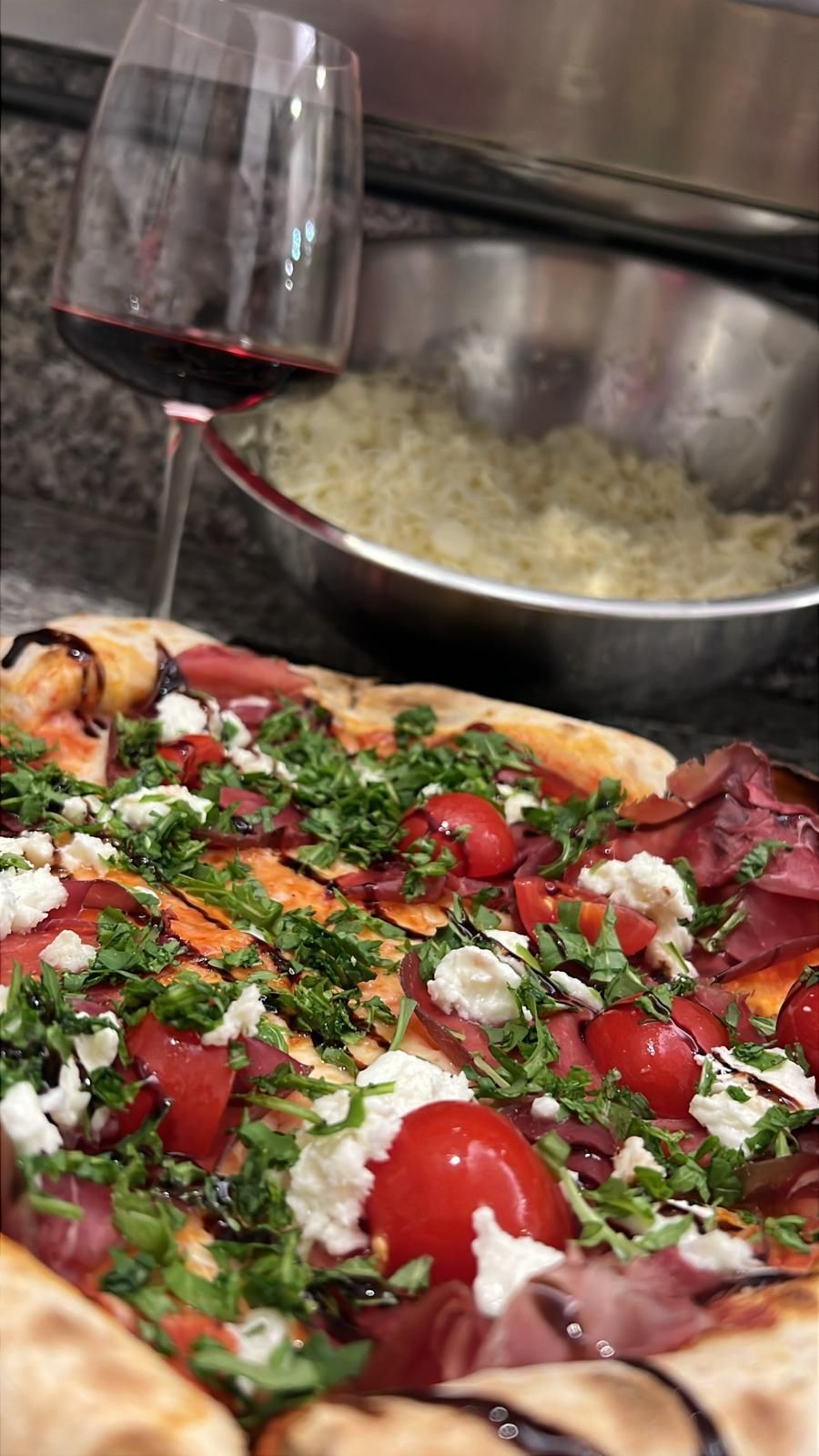 Pizza condita con pomodori, erbe aromatiche, formaggio e prosciutto; bicchiere di vino rosso.