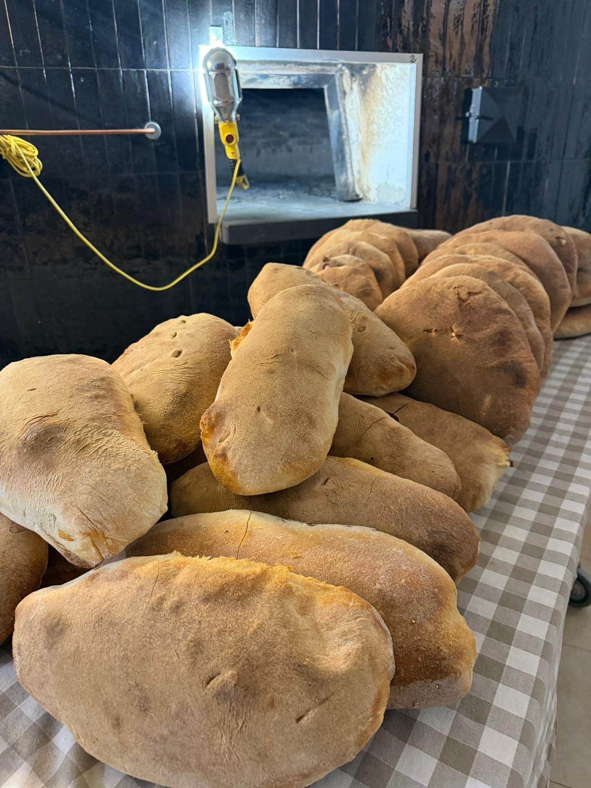 Pane pita appena sfornato e dorato, impilato su una tovaglia a quadretti vicino a un forno industriale.