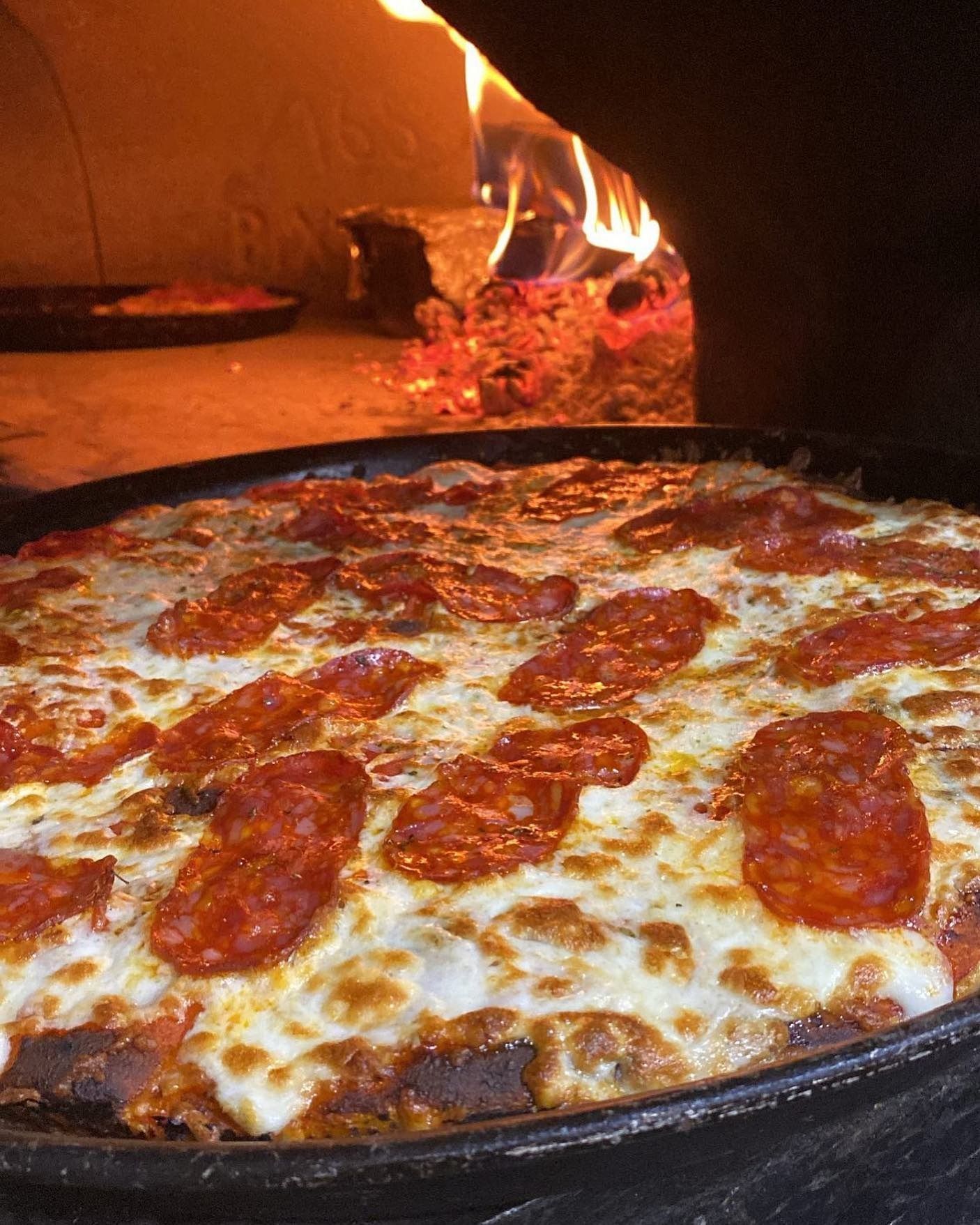 Pizza al pepperoni cotta nel forno a legna; fiamme e ceppi sullo sfondo.