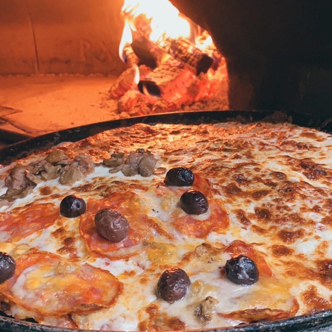 Pizza cotta nel forno a legna. Con formaggio, salame piccante, olive e funghi. Sullo sfondo, il fuoco arde.