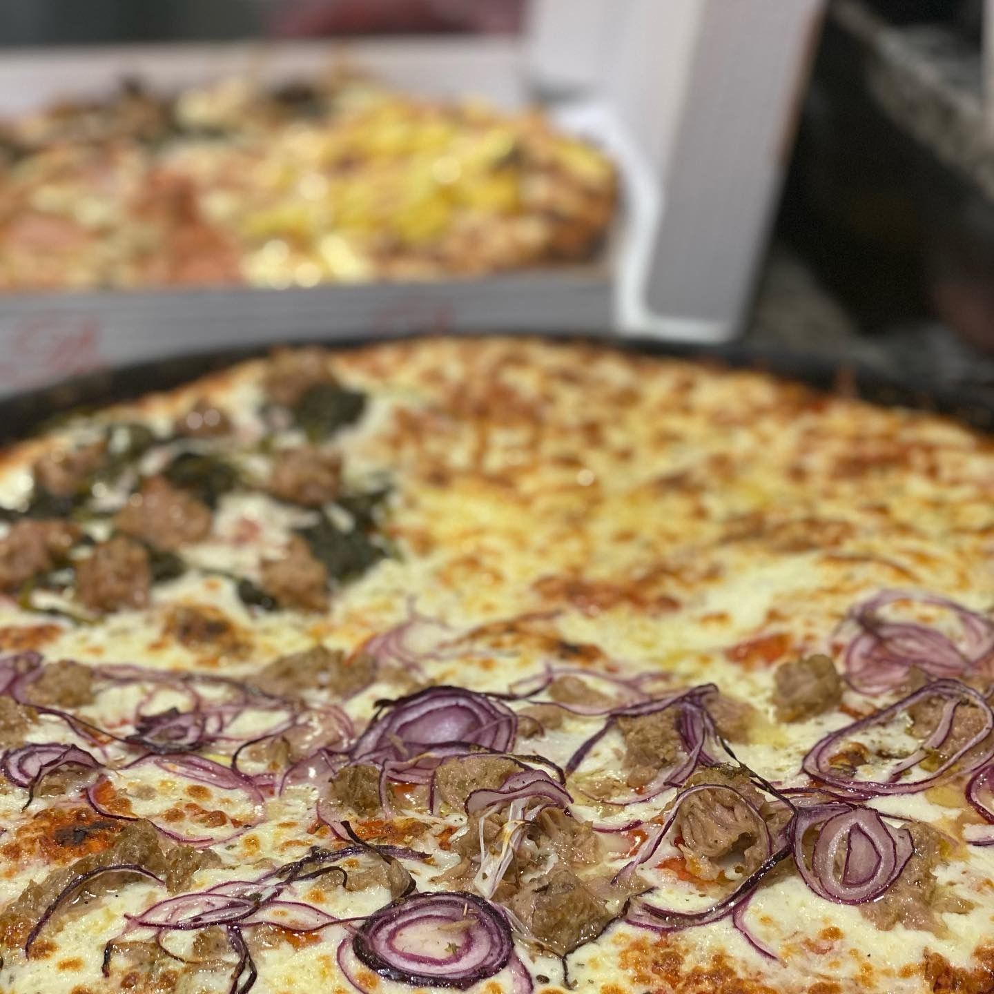 Primo piano di una pizza grande, divisa in sezioni con diversi condimenti: salsiccia, cipolla rossa e formaggio. Un'altra pizza è sfocata sullo sfondo.