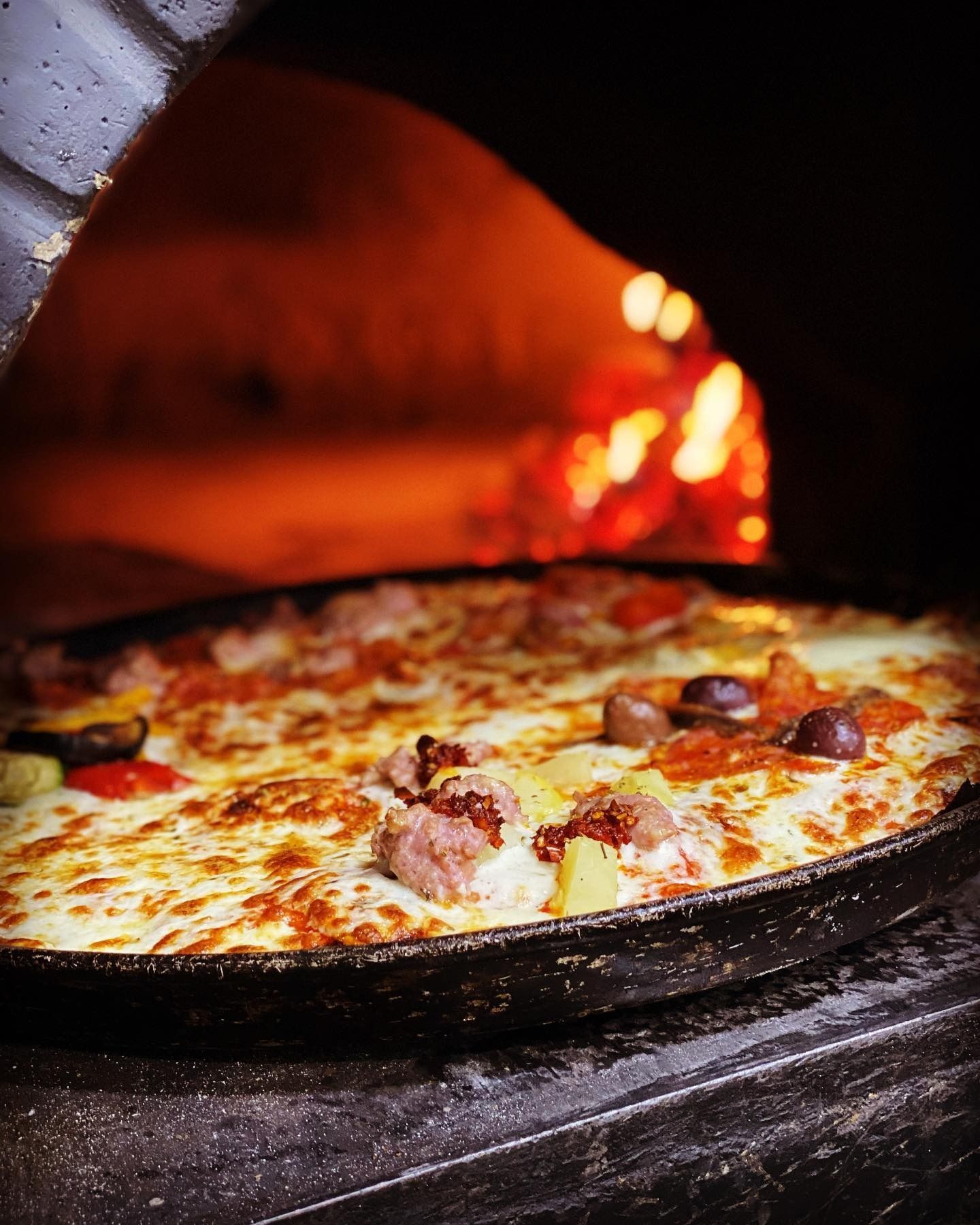 Pizza in un forno buio, con la crosta dorata, i condimenti visibili e un fuoco acceso sullo sfondo.