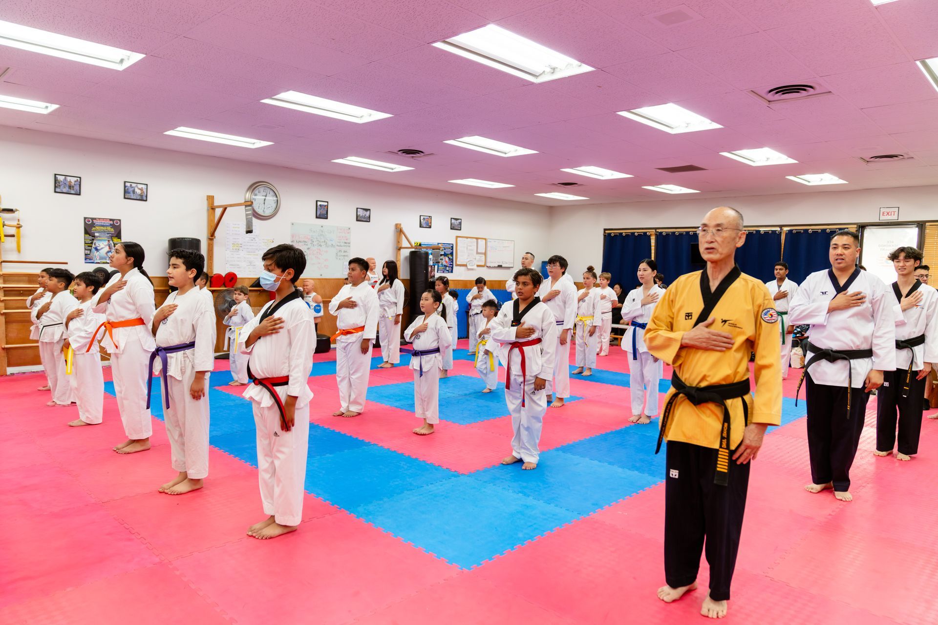 Taekwondo students in white uniforms practice poses in a studio with pink and blue floor mats and flags on the wall.