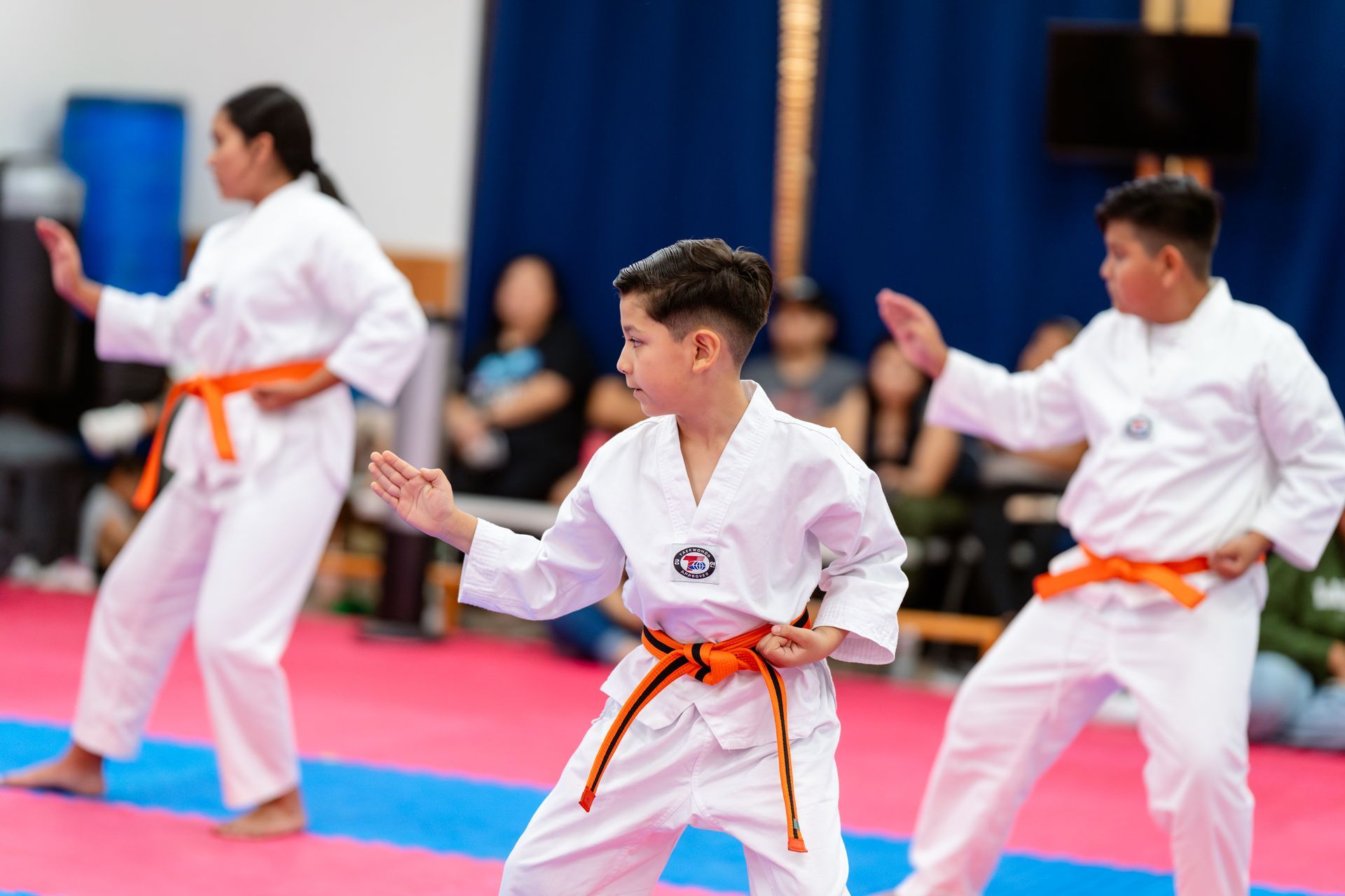 Two martial arts students in white uniforms practicing a defensive leg-block technique on a blue and pink mat.
