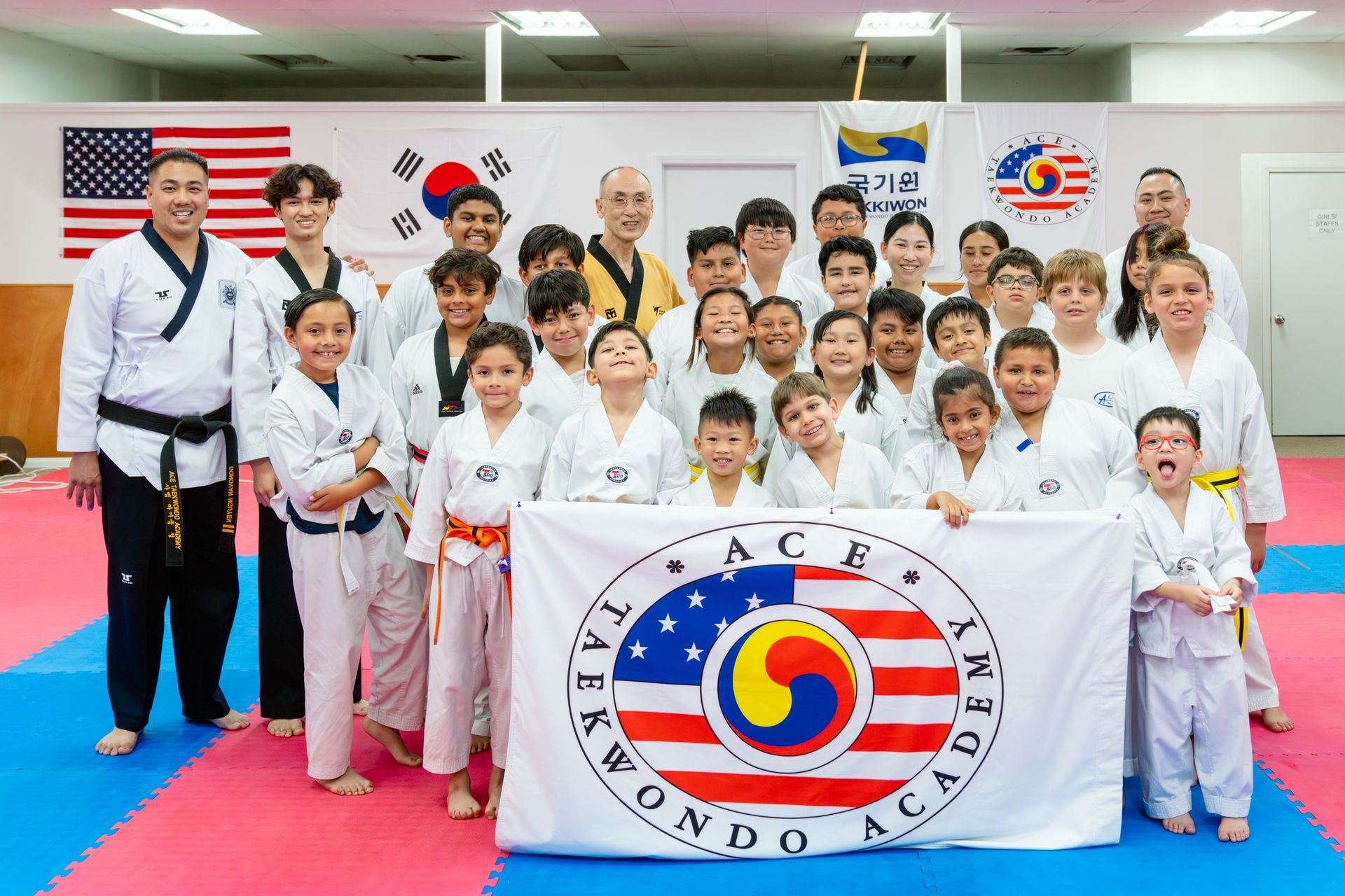 A group of taekwondo students and instructors pose with a branded academy flag in a training studio.
