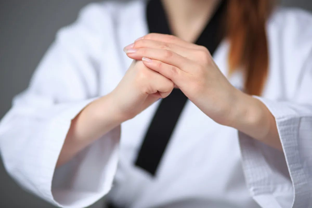 Person in a white martial arts uniform with a black collar performing a formal hand gesture against a gray background.