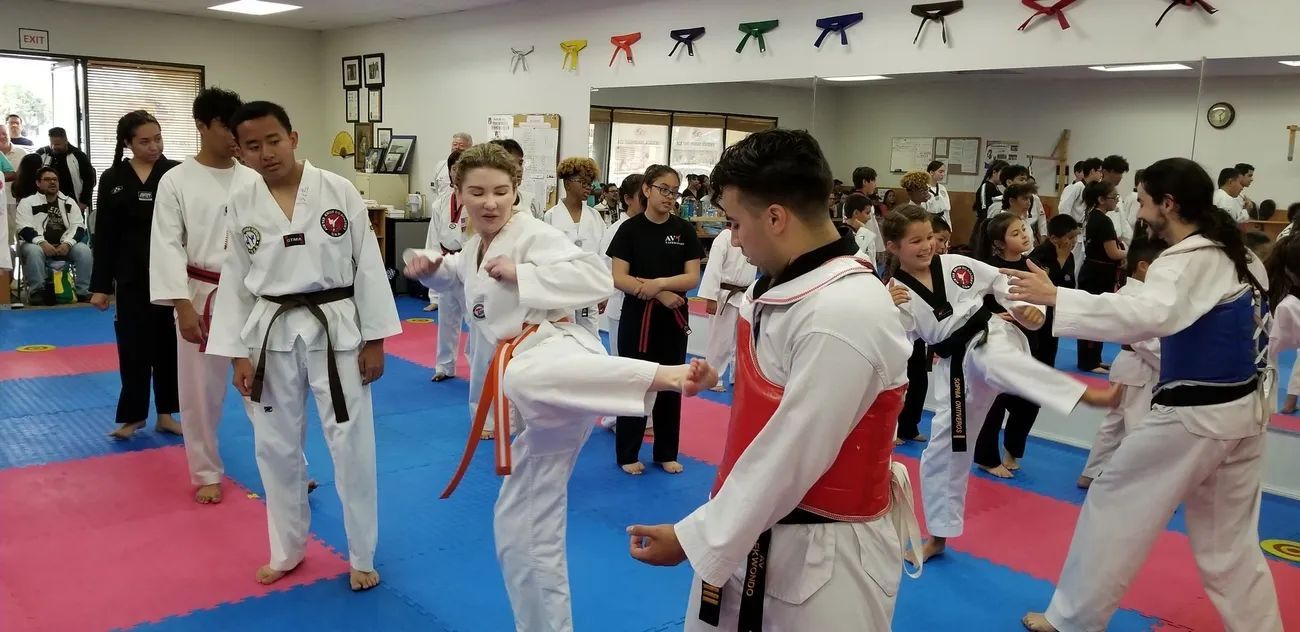 A taekwondo class in a studio with students in white uniforms practicing kicks with protective gear.