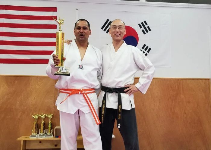 Two individuals in white martial arts uniforms pose in front of US and South Korean flags, holding a gold trophy.