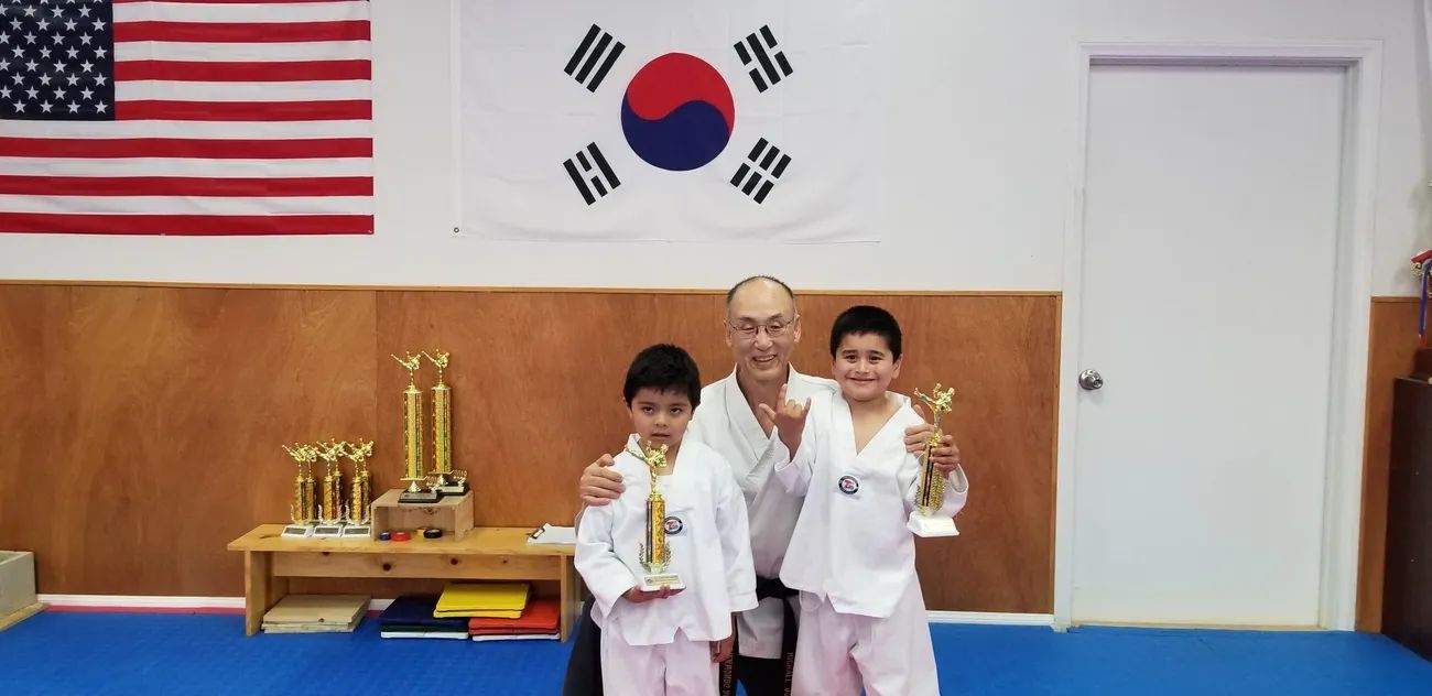 An instructor and two students in white martial arts uniforms hold gold trophies in a studio with American and Korean flags.