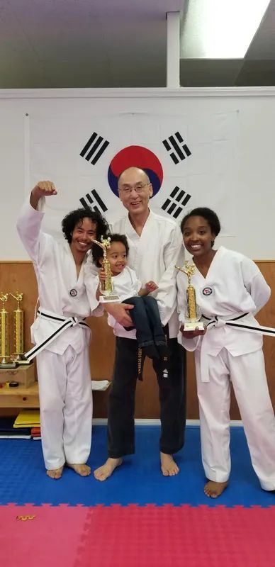 A group in white martial arts uniforms holding trophies stands in a training studio with a South Korean flag background.