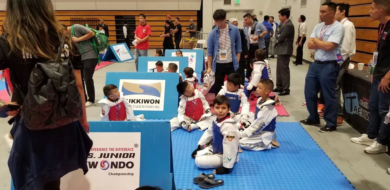Children in taekwondo uniforms sit on a blue mat at an indoor competition, surrounded by spectators and signage.