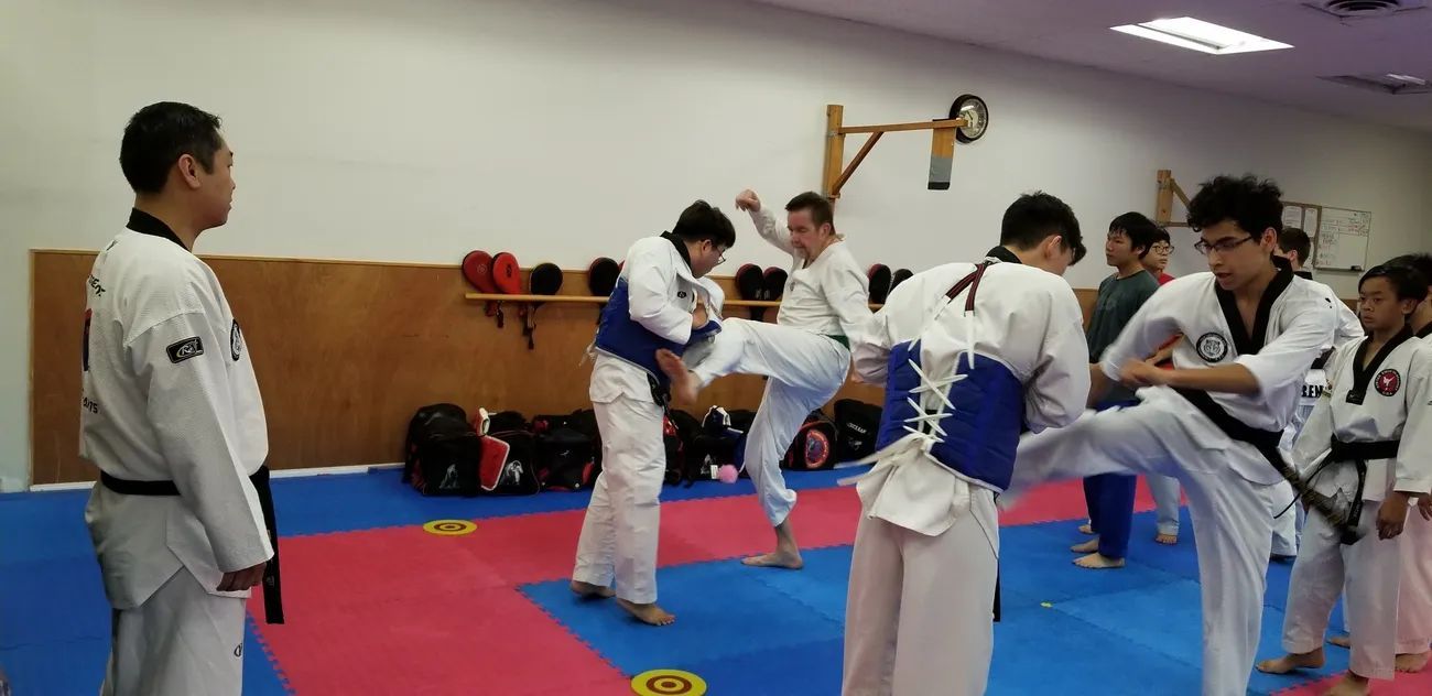 Taekwondo students practice kicking techniques in a gym, with some wearing white uniforms and blue protective gear.