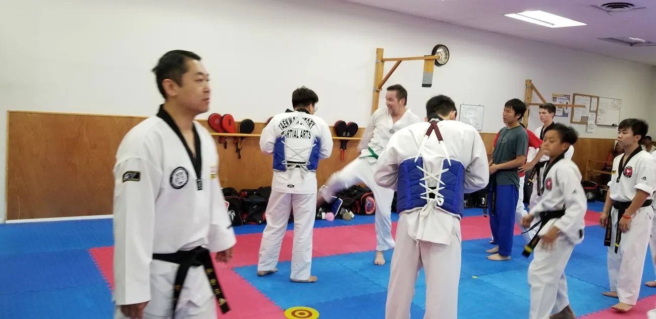 People in taekwondo uniforms practice sparring and drills in a gym with red and blue mats.