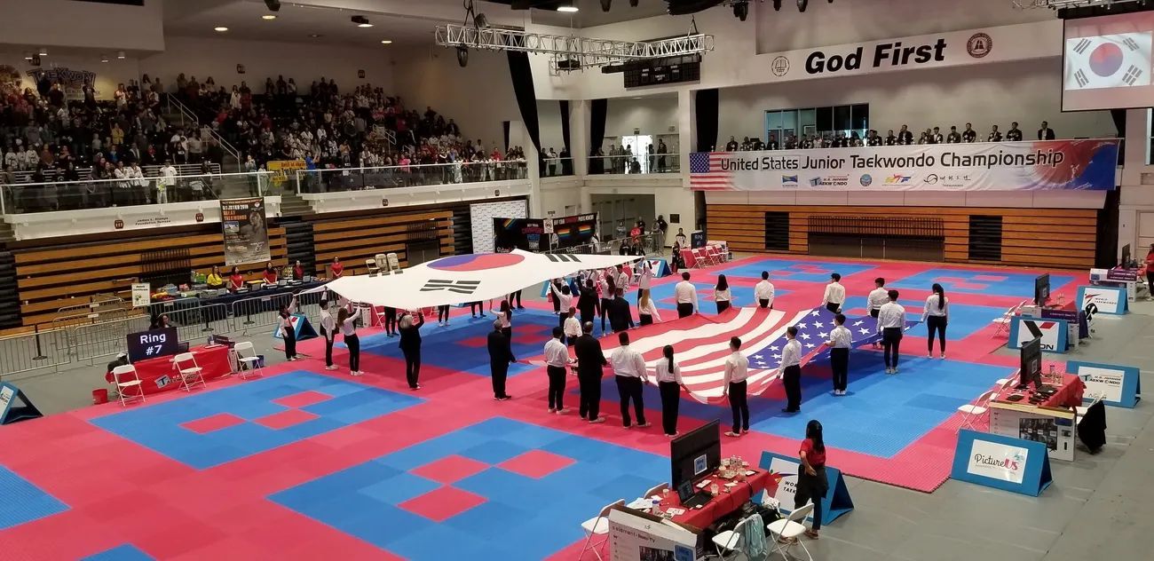 People in uniforms hold large South Korean and American flags in the center of an indoor martial arts arena.