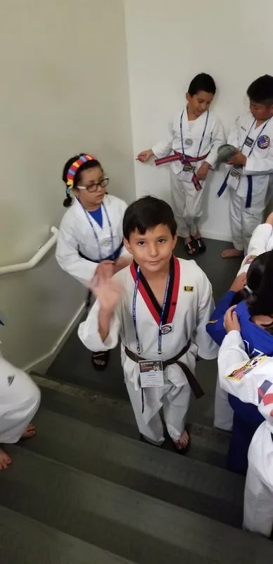 A group of children in white karate uniforms stands on a staircase; one child in the center waves at the camera.