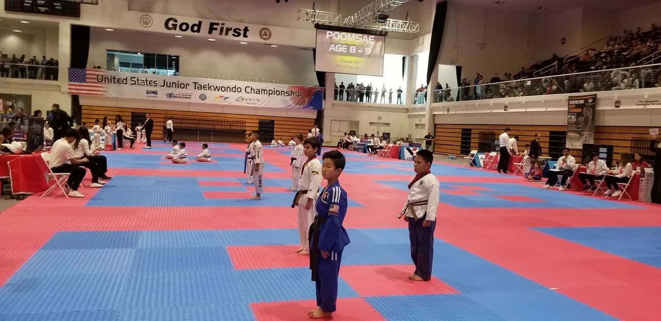Children in martial arts uniforms stand on a red and blue padded mat in a large indoor competition venue.