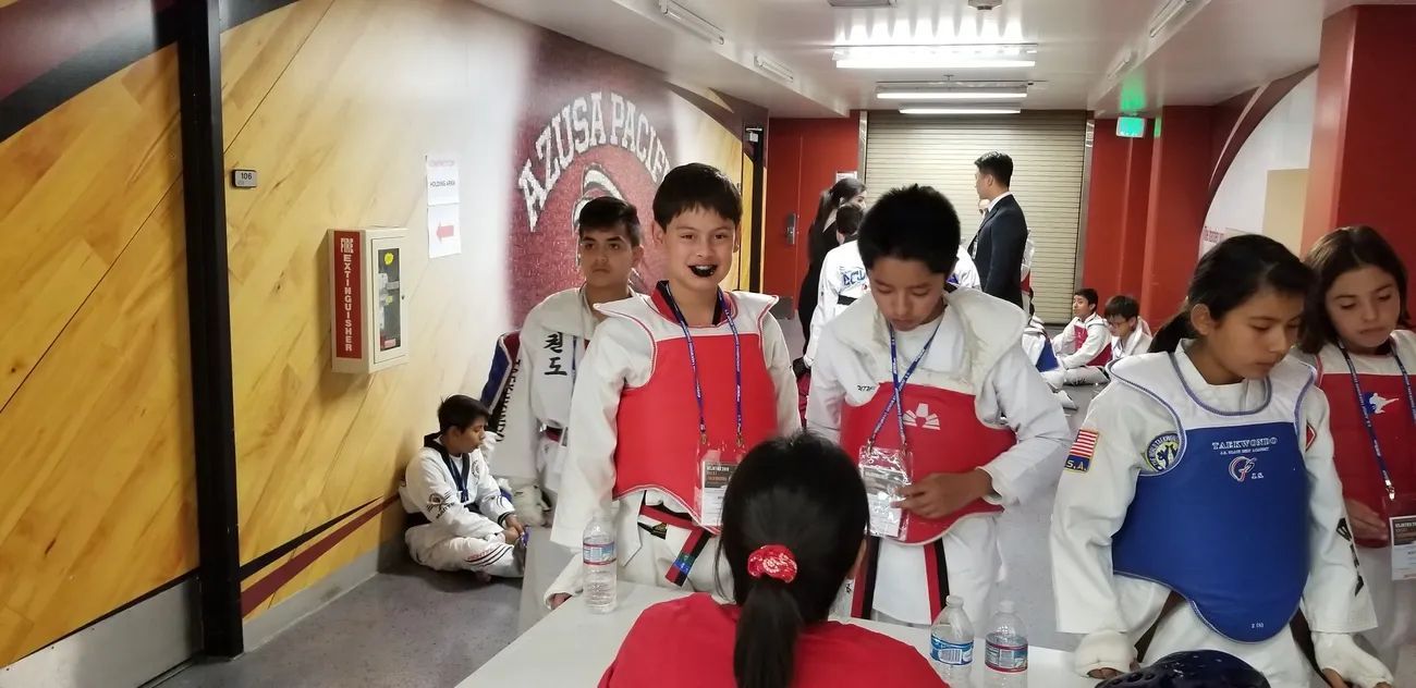 Taekwondo participants in protective gear stand in a hall near a wall with a logo, with one sitting in the background.