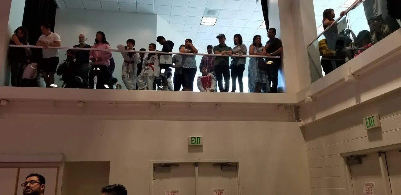Spectators standing on an indoor balcony watching an event from above in a brightly lit room with exit signs.