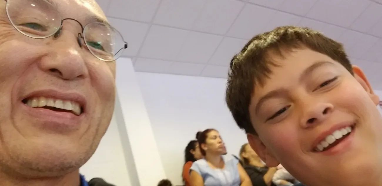 Close-up selfie of a smiling older man with glasses and a younger boy, with people blurred in the background.