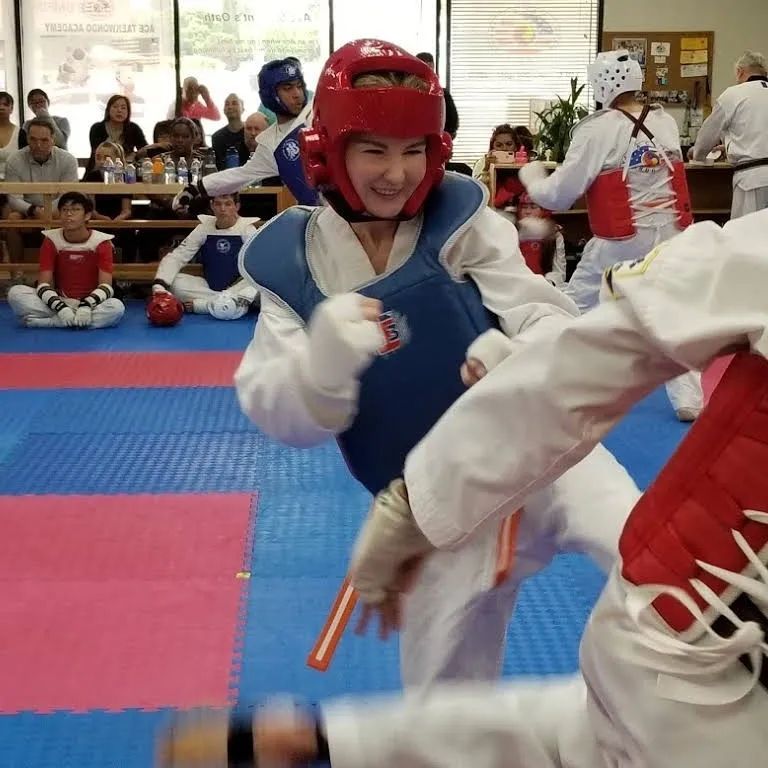 A sparring match in a taekwondo studio with participants wearing white uniforms and protective head and chest gear.