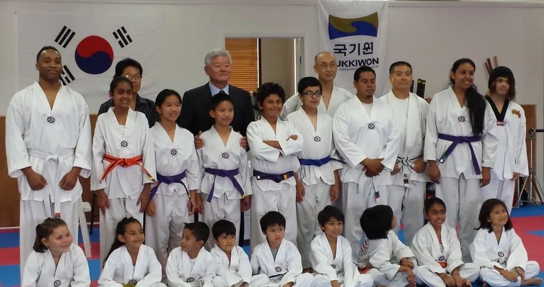Group of martial artists in white uniforms with colored belts pose with instructors in a dojang with a Korean flag.