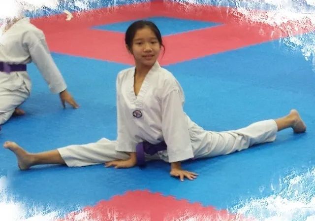 A person in a white taekwondo uniform with a purple belt performs a side split on a blue and red matted floor.