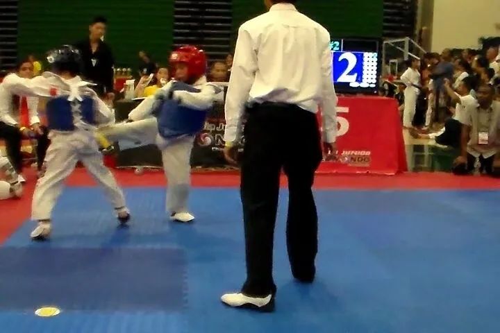 Two taekwondo athletes in white uniforms and sparring gear compete on a blue mat, observed by a referee in a white shirt.