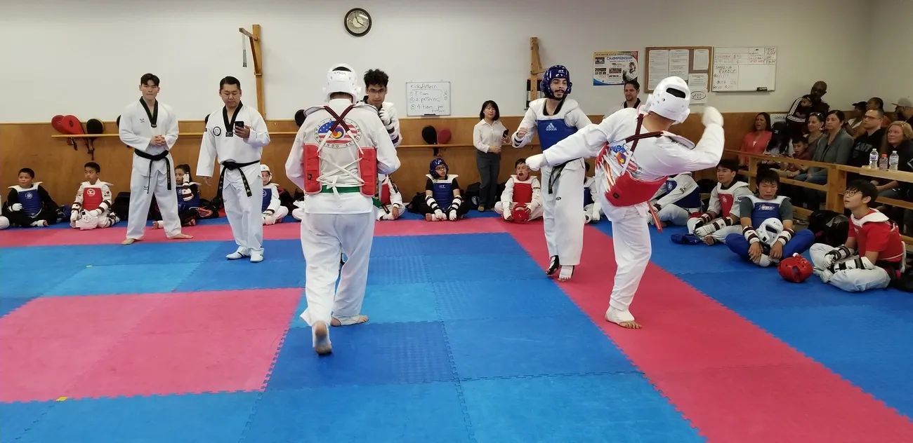 Taekwondo practitioners spar on blue and red mats in a training facility while others watch from the sidelines.