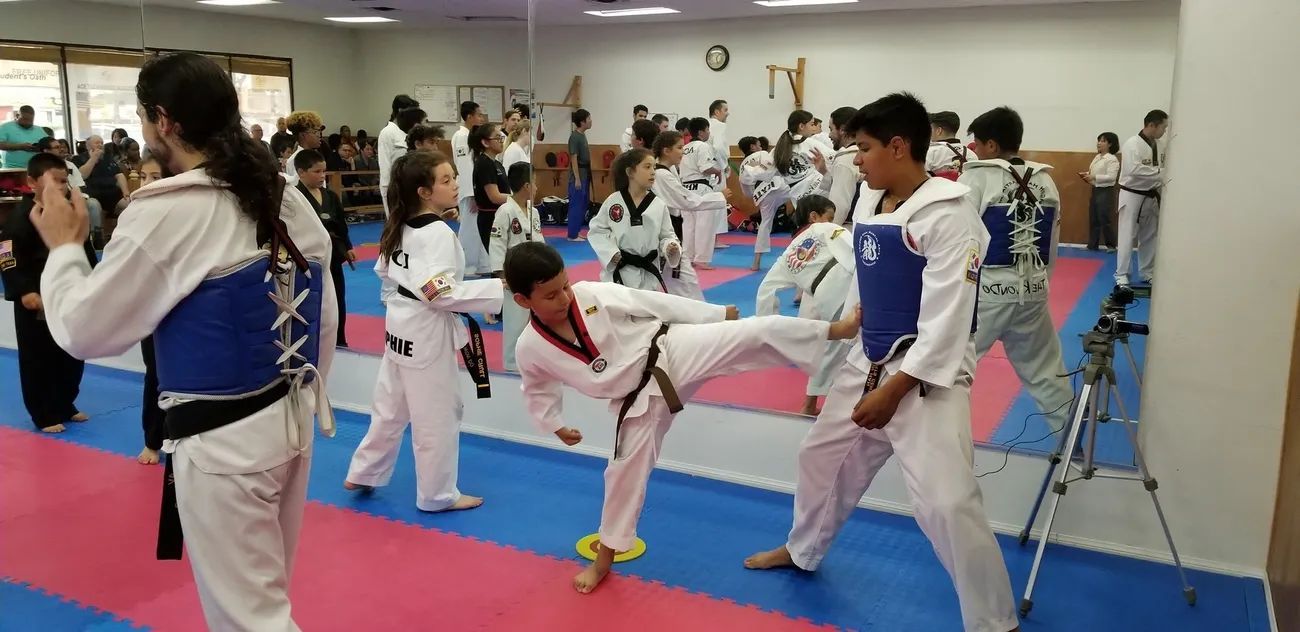 Students in taekwondo uniforms and blue chest protectors practice sparring drills inside a martial arts training gym.