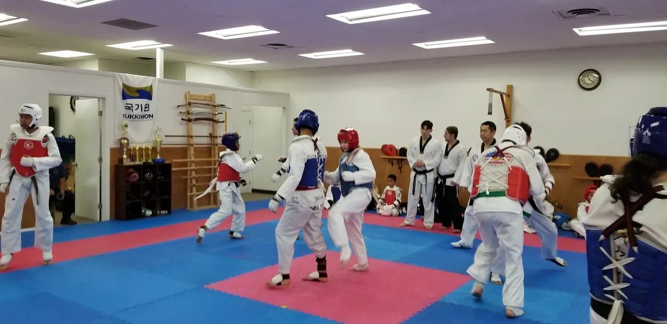 People in taekwondo uniforms and protective gear spar on a blue and pink mat in a training studio.