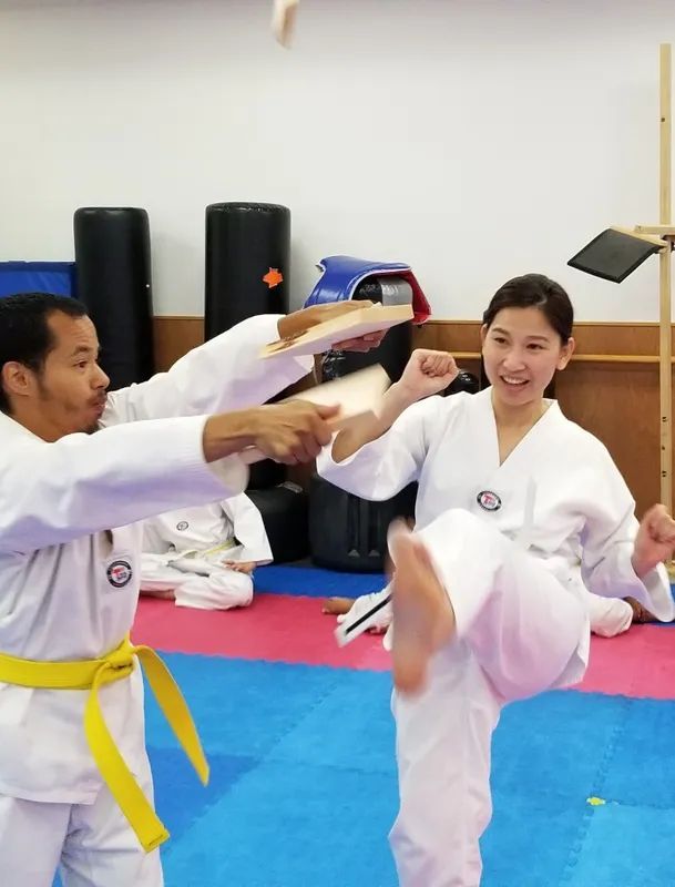 A person in a white uniform holding a wooden board for another person to practice a kick during a martial arts class.