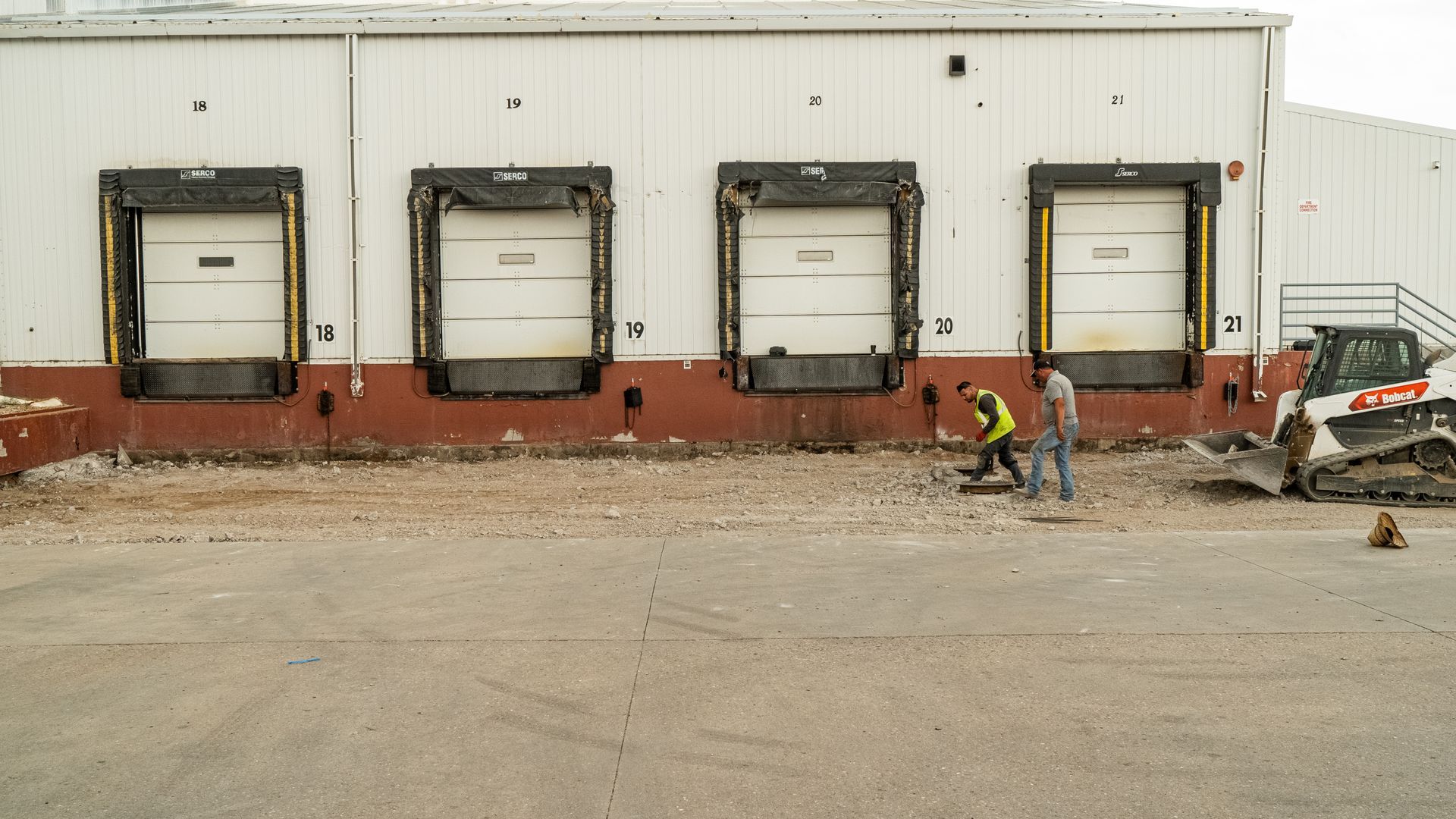 Workers and a skid steer beside four loading dock doors on a warehouse exterior.